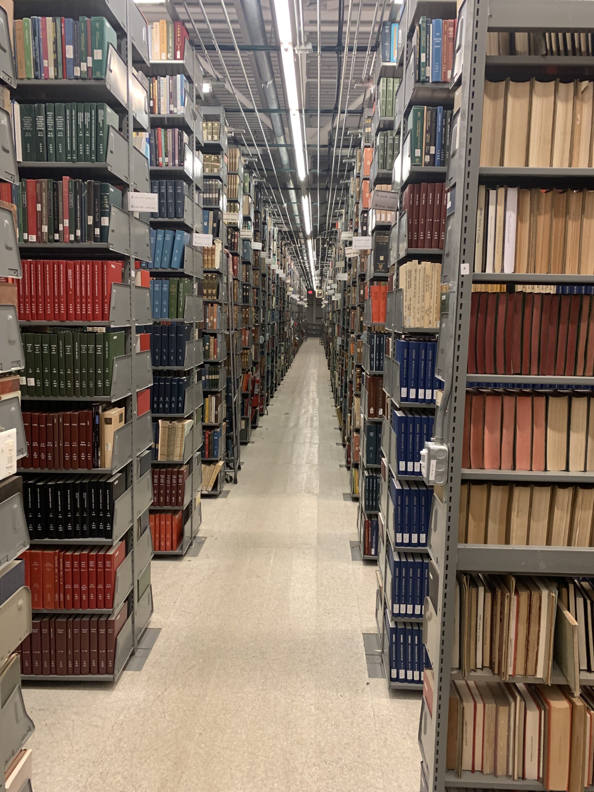 Rows of metal bookshelves filled with books and periodicals line both sides of a well-lit library aisle with a reflective tile floor.