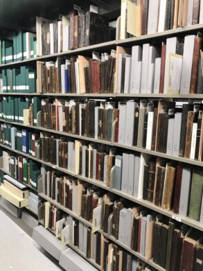 Shelves filled with old books and archival boxes organized in a library or archive storage area.