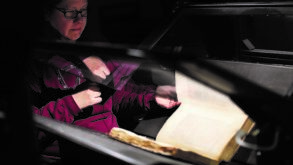 A person sits at a desk turning the pages of an old large book under a protective glass enclosure, with dim lighting focused on the book.