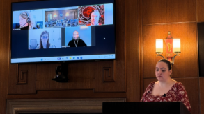 A woman stands at a podium in a wood-paneled room while a video conference with five participants is displayed on a large screen behind her.