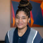 A person with glasses and dreadlocks styled in a bun, wearing a collared shirt and sweater, stands in front of a colorful geometric artwork.