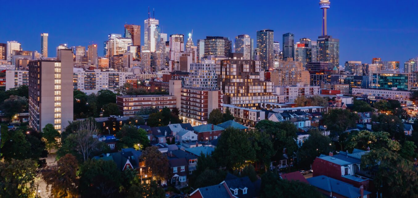 Toronto skyline at dusk, featuring the CN Tower and high-rise buildings above a residential neighborhood with trees and houses in the foreground.