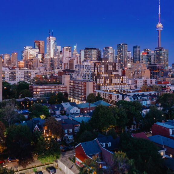 Toronto skyline at dusk, featuring the CN Tower and high-rise buildings above a residential neighborhood with trees and houses in the foreground.