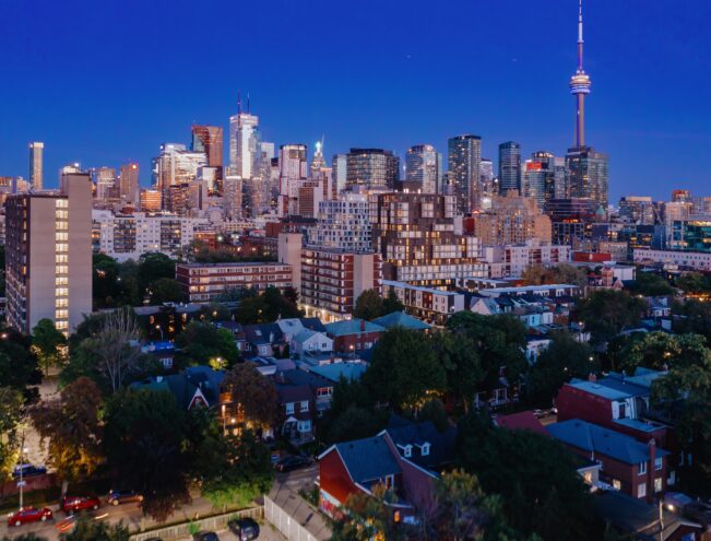 Toronto skyline at dusk, featuring the CN Tower and high-rise buildings above a residential neighborhood with trees and houses in the foreground.