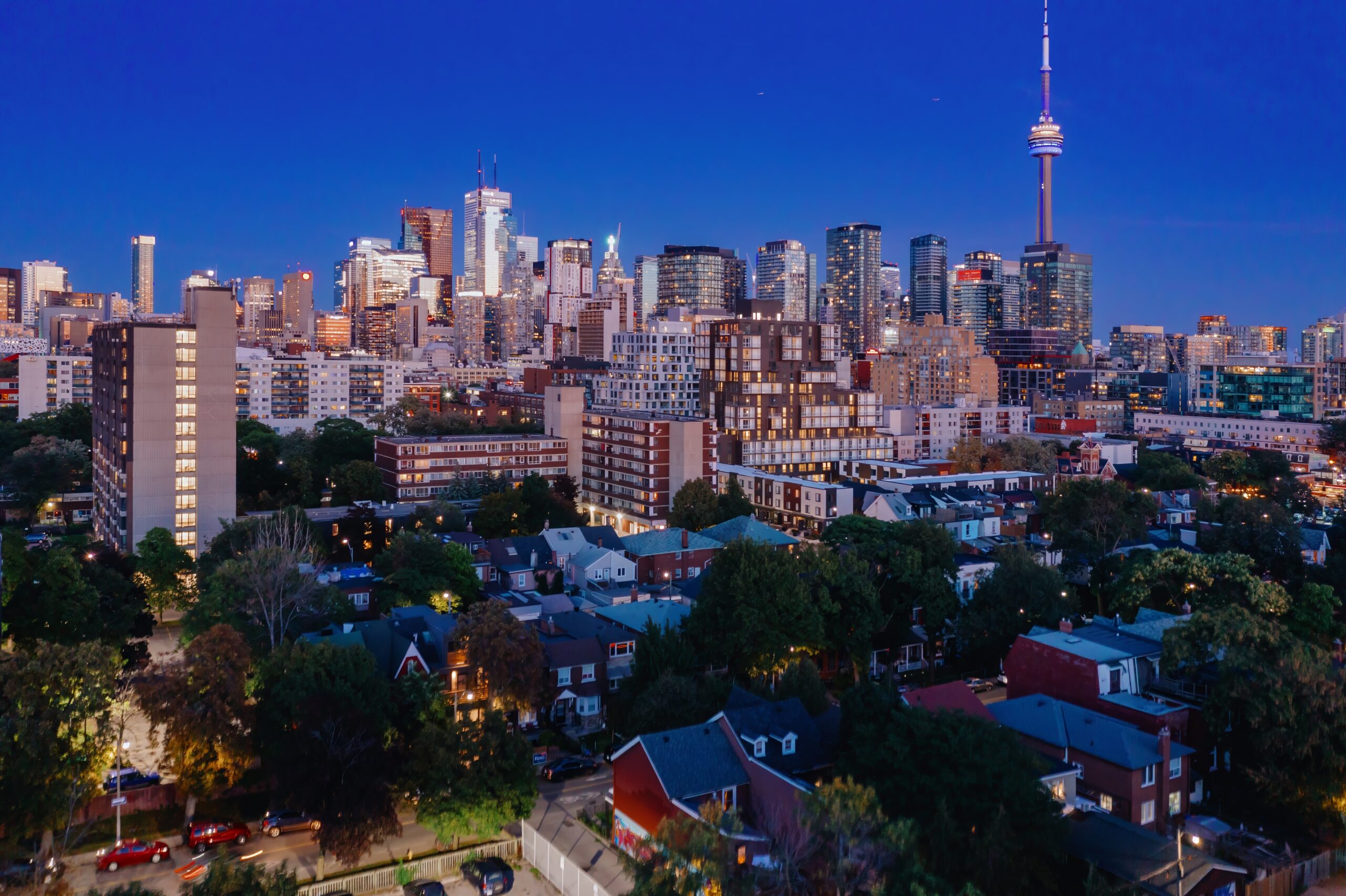 Toronto skyline at dusk, featuring the CN Tower and high-rise buildings above a residential neighborhood with trees and houses in the foreground.