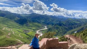 Person in a blue jacket and sunhat stands on a stone path overlooking a green valley with winding roads, river, and mountains under a partly cloudy sky.