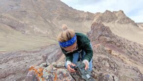 Person wearing a blue "Colby" headband examines a camera on rocky terrain with mountains in the background.