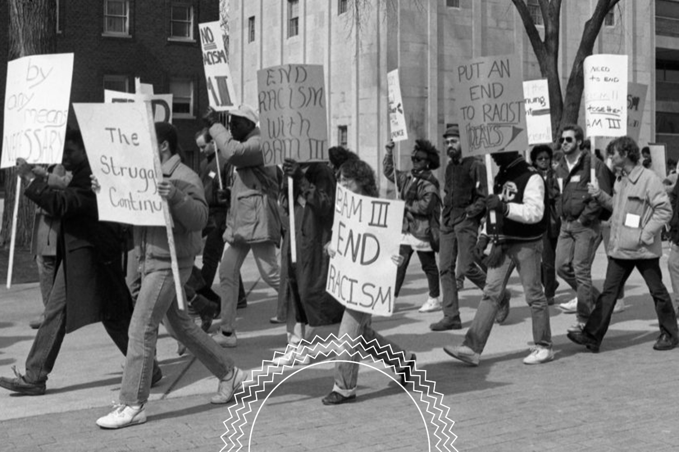 A group of people march outdoors holding signs with messages opposing racism, such as “End Racism” and “Put an End to Racist Tradism.”.