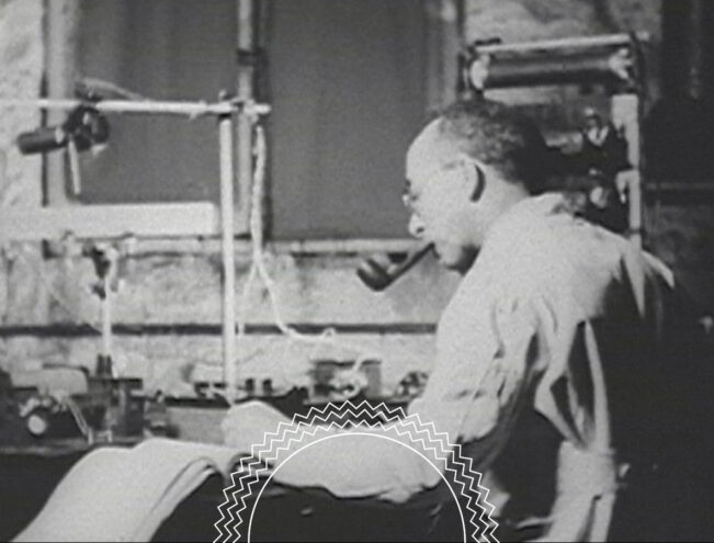A man with a pipe works at a desk covered with papers and scientific equipment in a black and white photo.