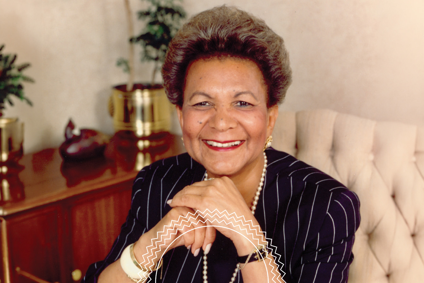 A woman in a striped suit sits in an office chair, smiling with hands folded. There are potted plants and a wooden cabinet in the background.