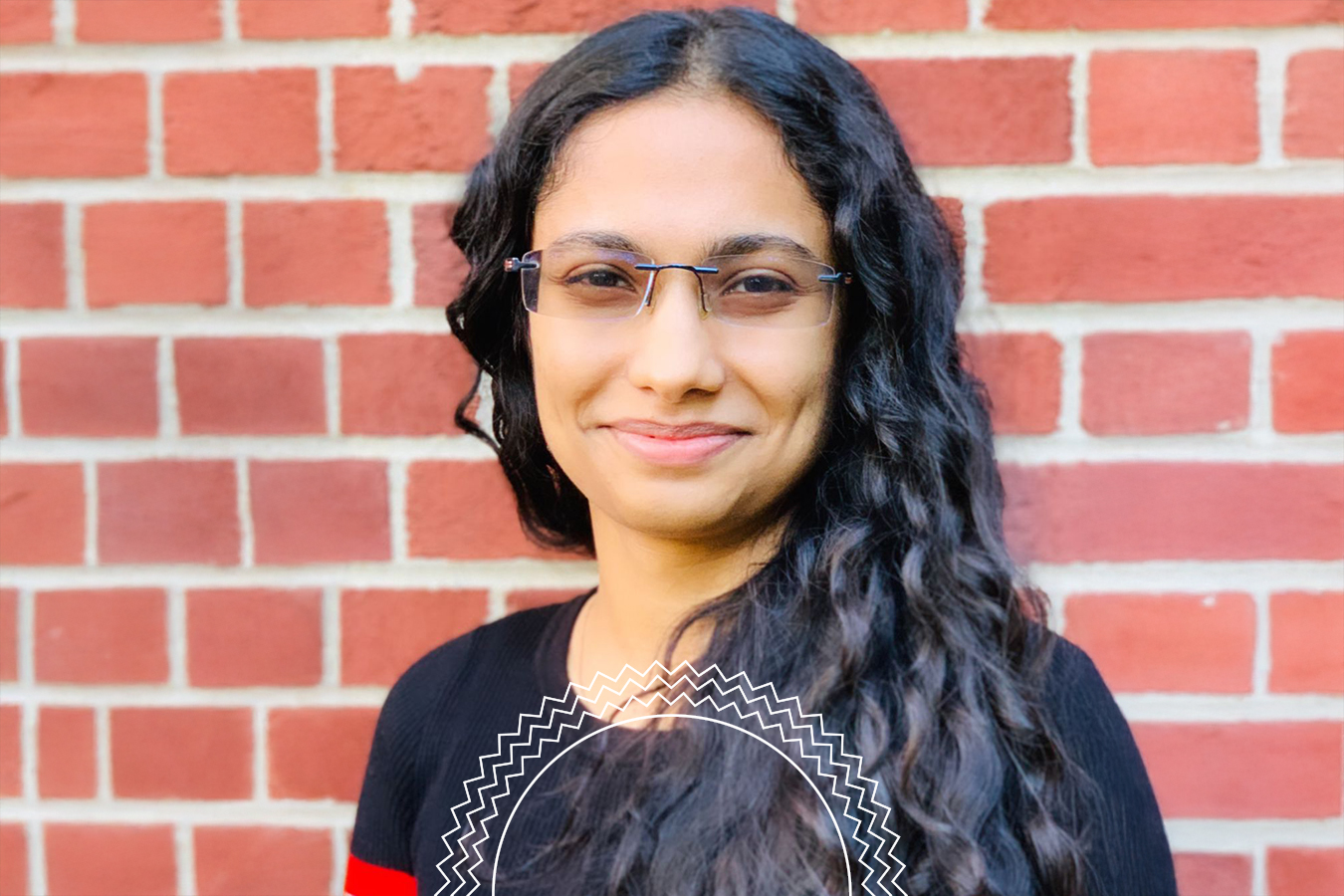 A person with long, wavy dark hair and glasses is smiling in front of a red brick wall, wearing a black and red top.