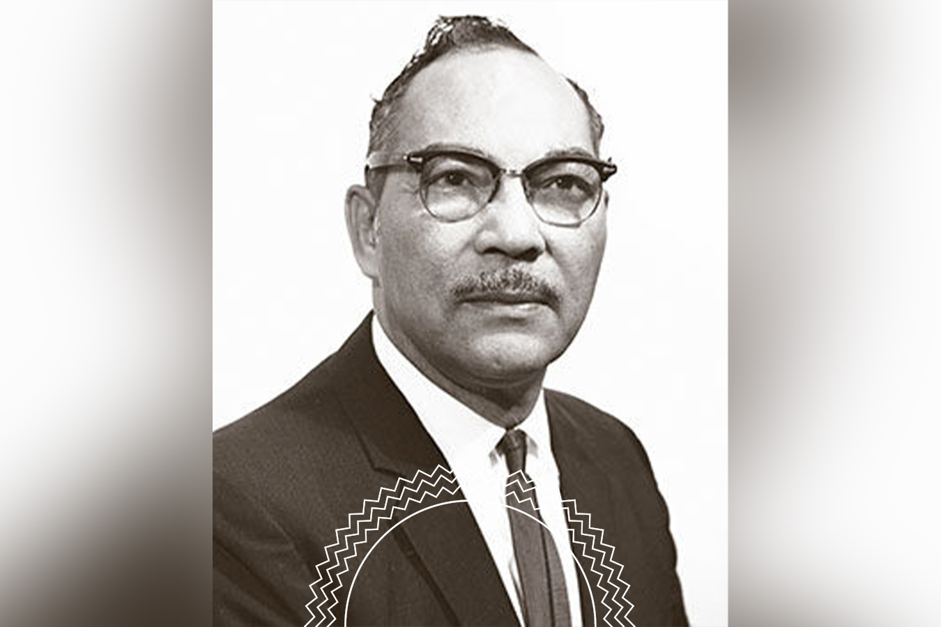 Black-and-white portrait of a man wearing glasses, a suit, and a tie, looking directly at the camera with a neutral expression.