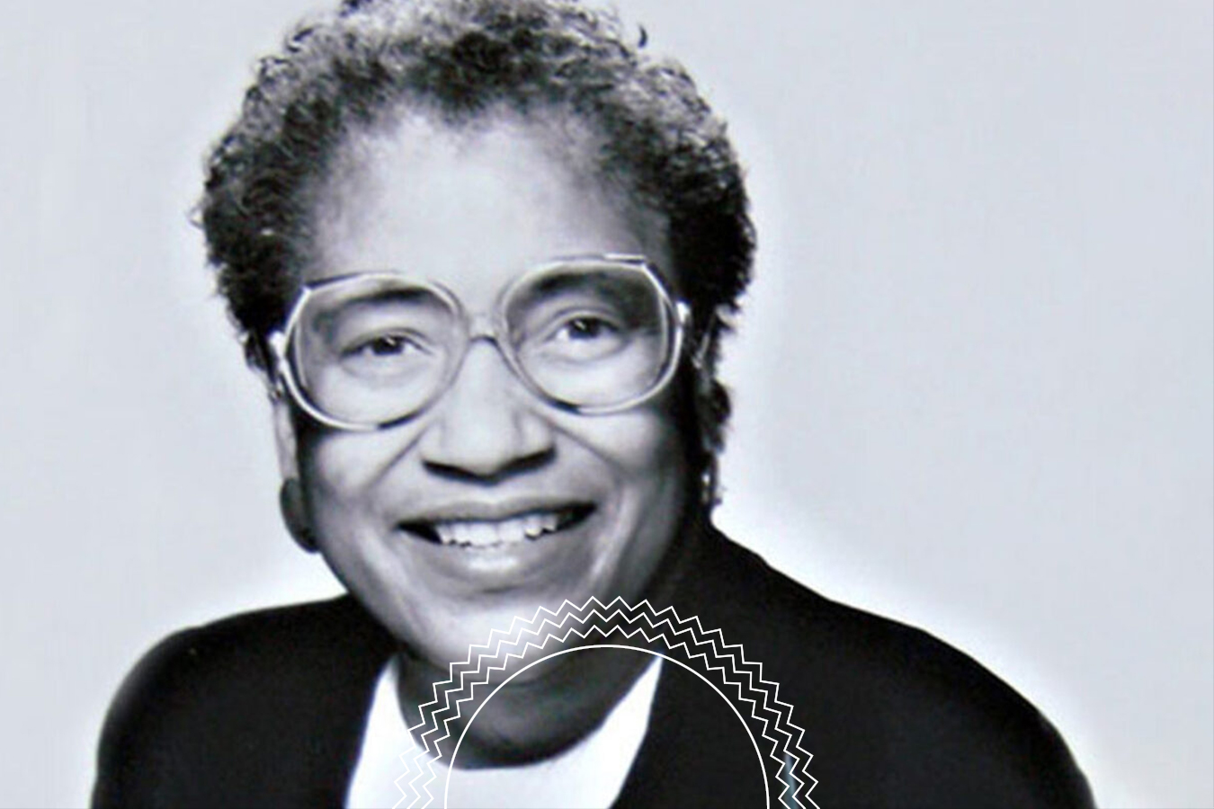 Black-and-white portrait of an older woman with short curly hair, glasses, and a suit jacket, smiling at the camera.