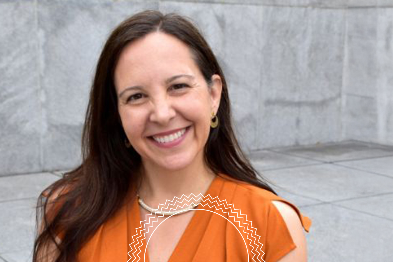 A woman with long brown hair, wearing an orange sleeveless top and gold earrings, smiles while standing in front of a gray stone wall.
