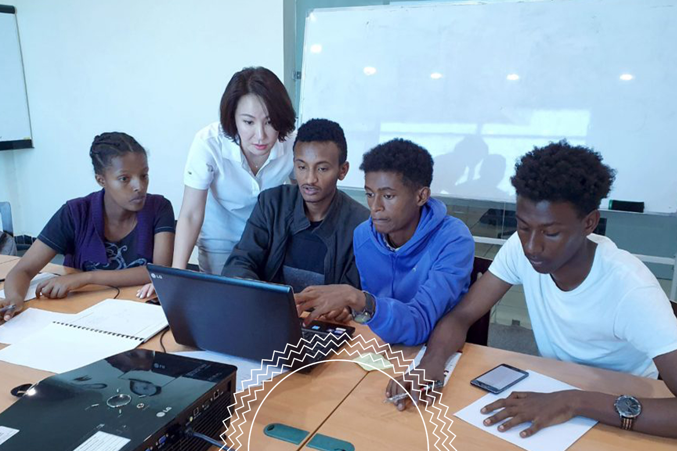 Five people sit around a table with a laptop and papers, collaborating in a classroom with a whiteboard in the background.