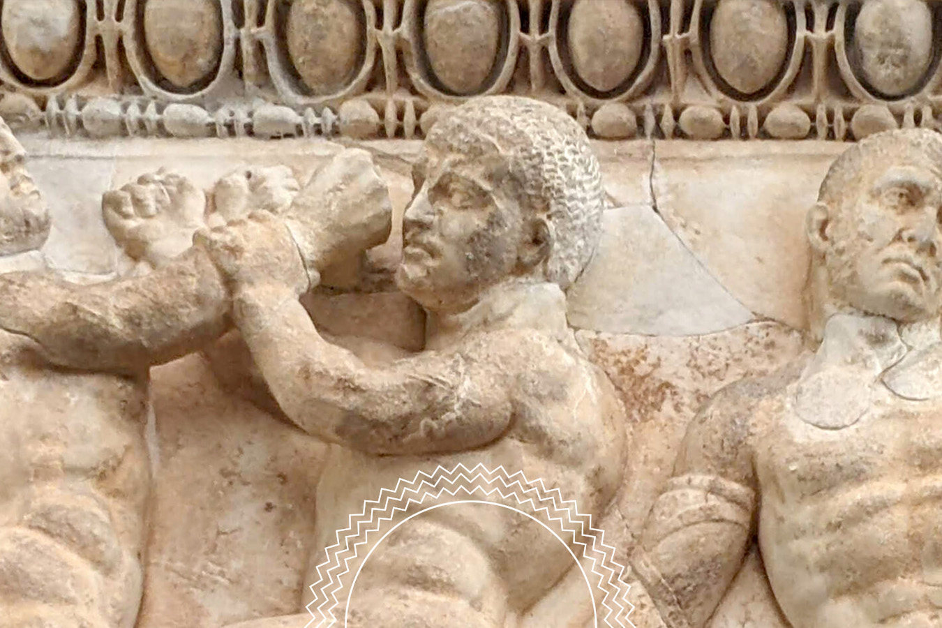 Close-up of an ancient stone relief showing three muscular male figures, with one figure appearing to block or push away another’s fist. Ornate carvings decorate the top edge.