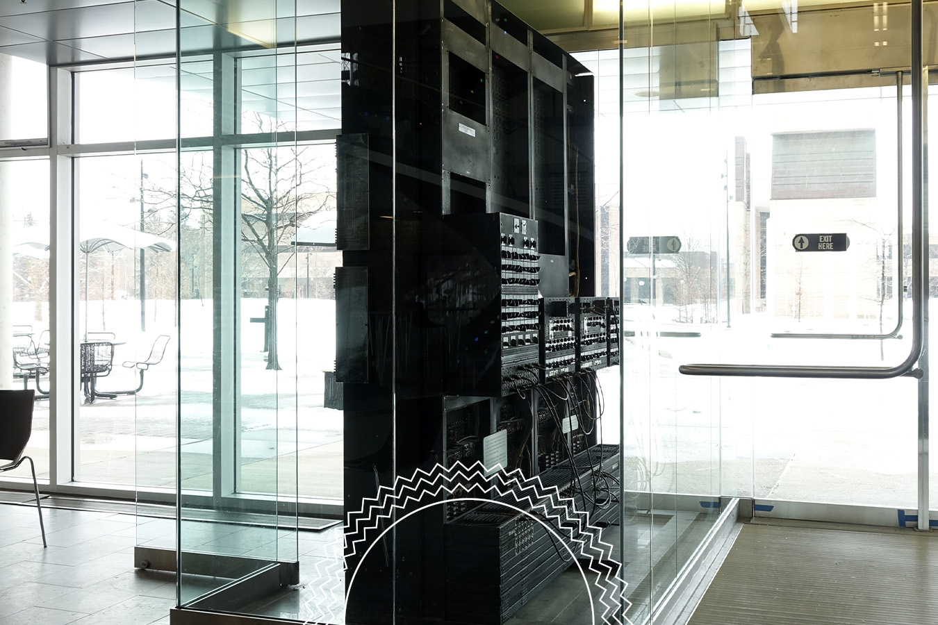 A black server rack stands inside a glass enclosure in a modern building lobby with large windows showing a snowy outdoor scene.