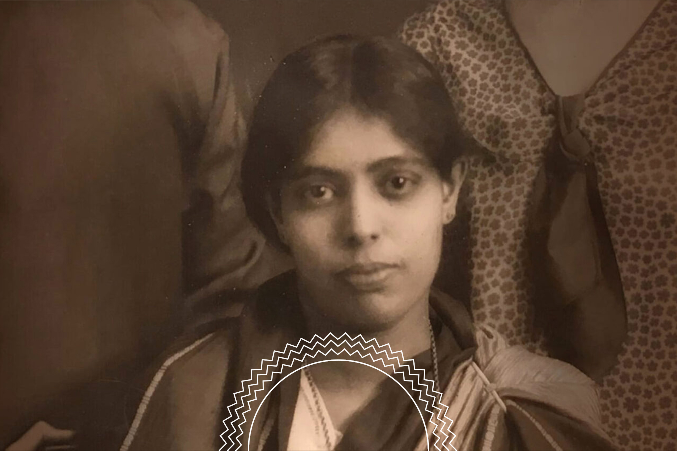 A sepia-toned portrait of a woman with short hair, wearing a patterned blouse and shawl, looking directly at the camera. Two partially visible people are in the background.