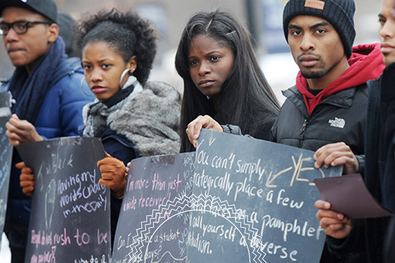 Four people stand outdoors holding handwritten signs, wearing winter clothing. They face forward with serious expressions.