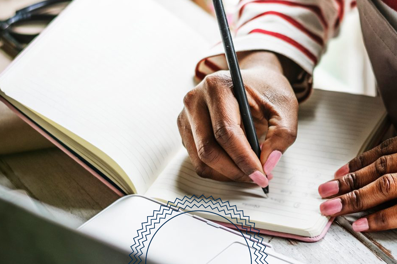 A person writes with a pencil in a lined notebook next to a laptop on a wooden table.