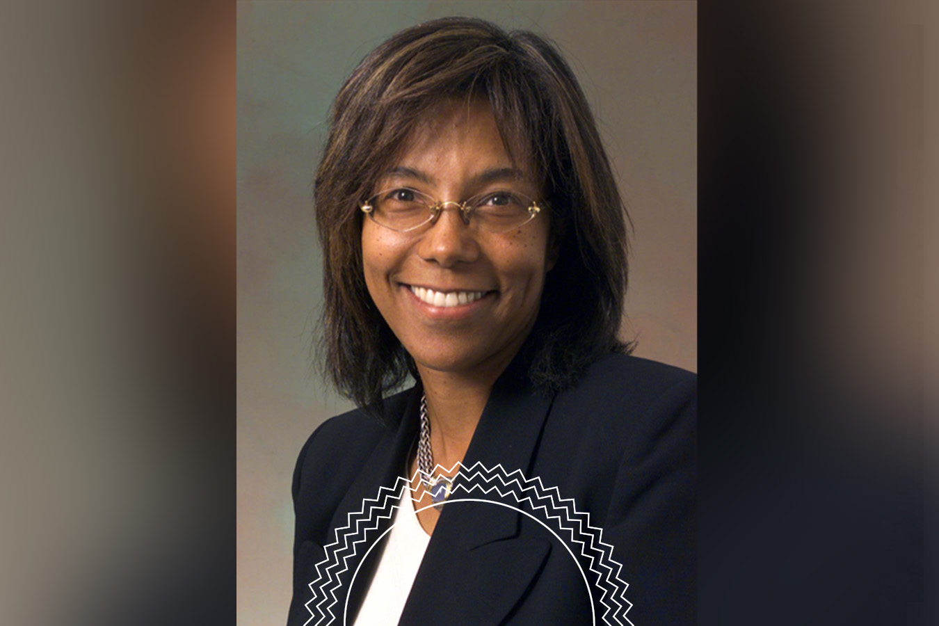 A woman with shoulder-length dark hair and glasses, wearing a black blazer over a white top, smiles at the camera against a neutral background.