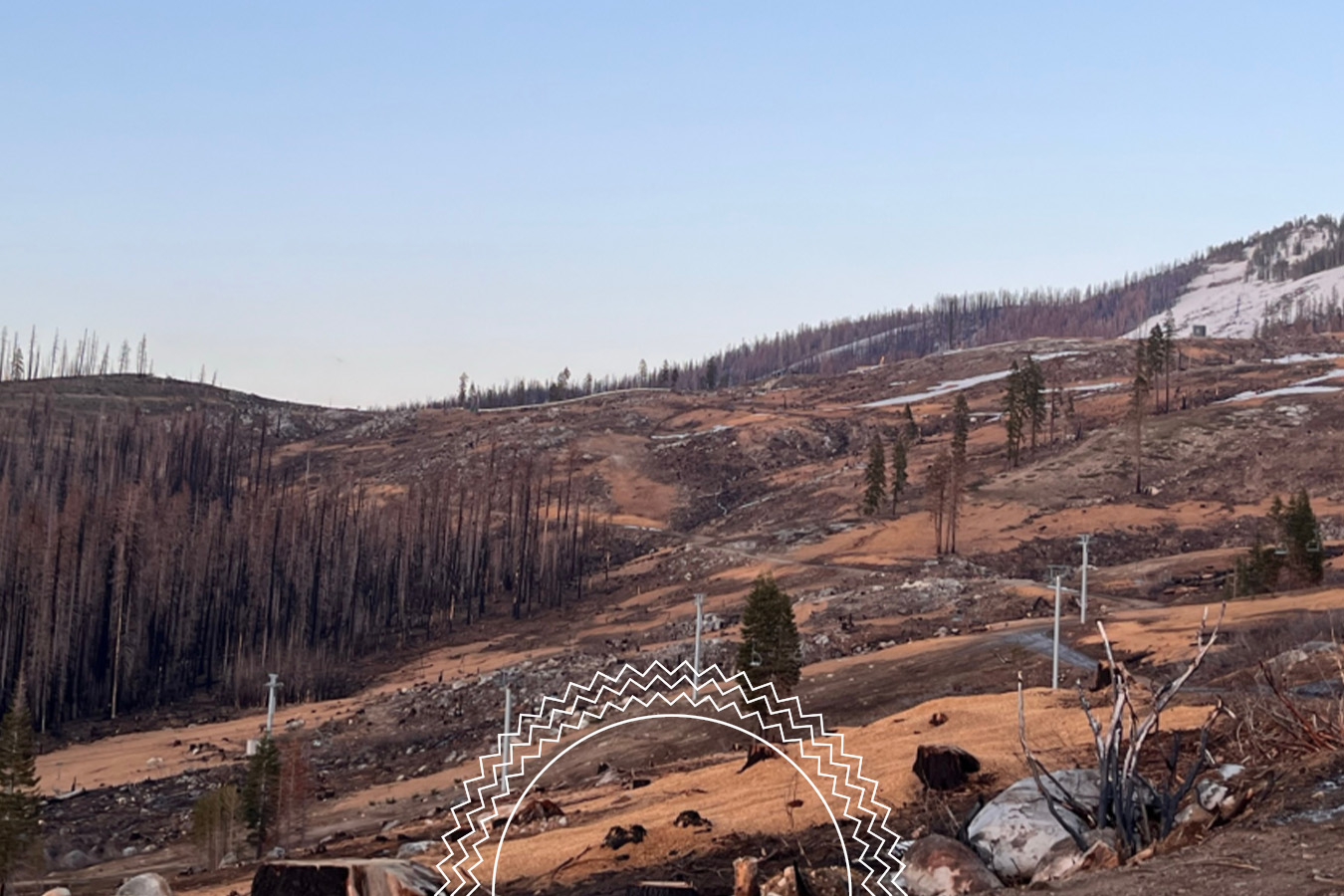 A barren, hilly landscape with scattered trees, patches of snow, and evidence of recent wildfire damage under a clear sky.