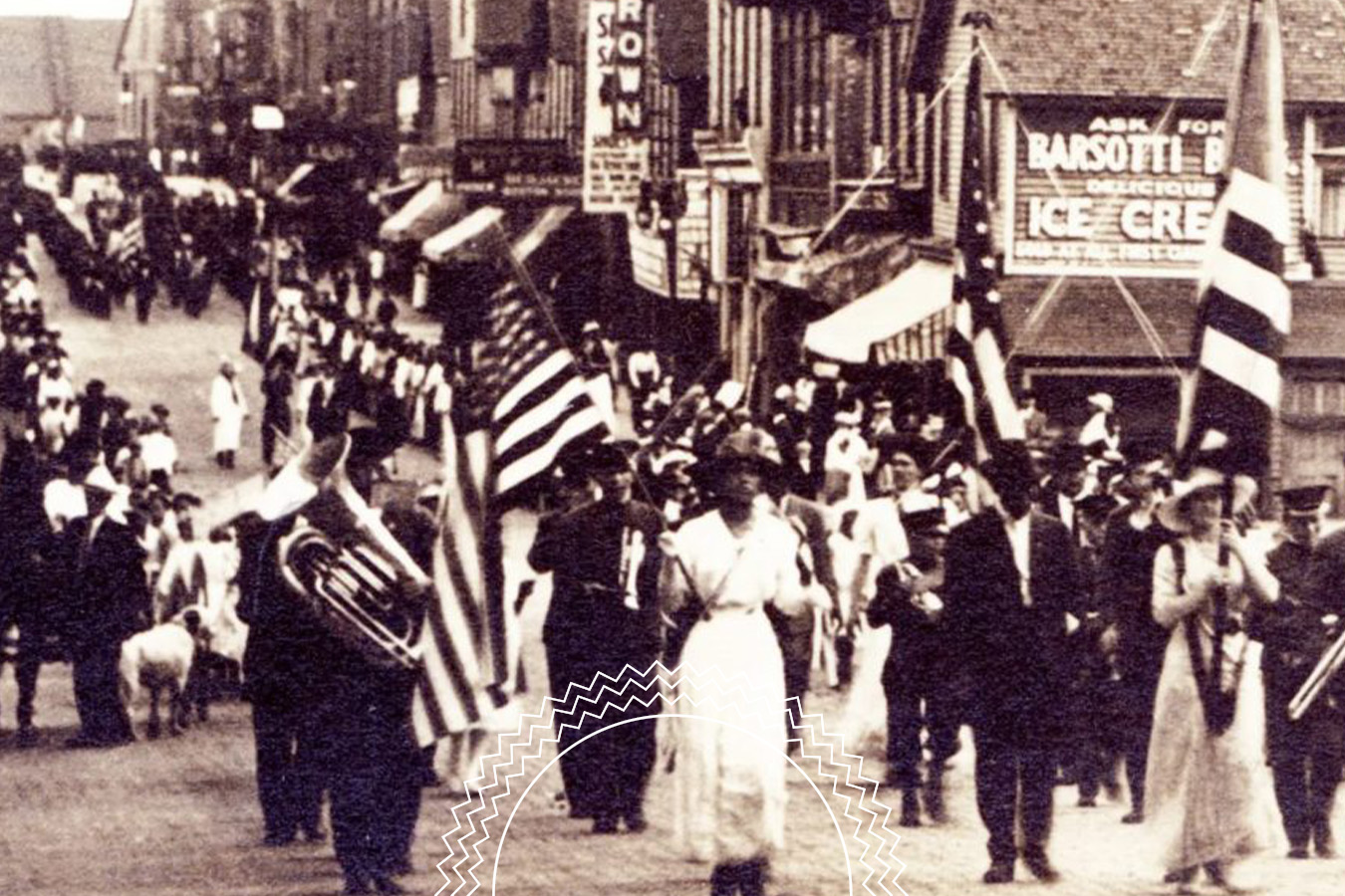A black and white photo of a parade on a busy street, featuring people marching with American flags and musicians, with storefronts in the background.