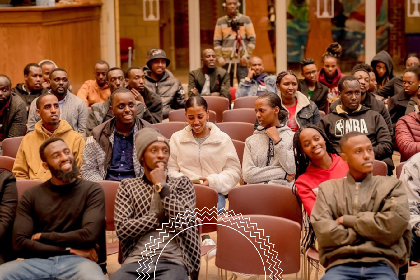 A group of people sit in rows of chairs, attentively watching an event indoors. Most are dressed warmly and appear engaged or smiling.