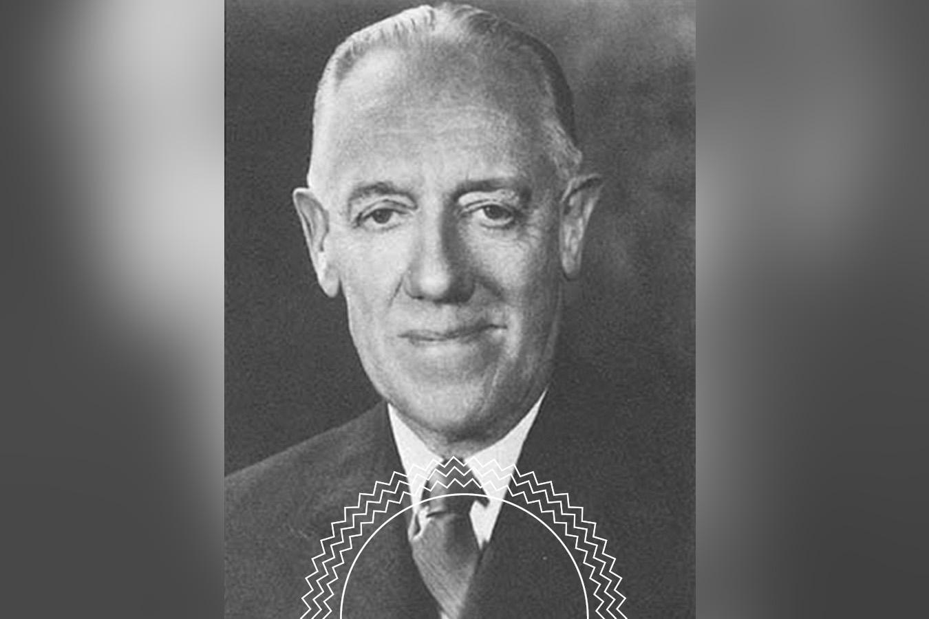 Black and white portrait of an older man in a suit and tie, facing the camera and smiling slightly, with a blurred background.