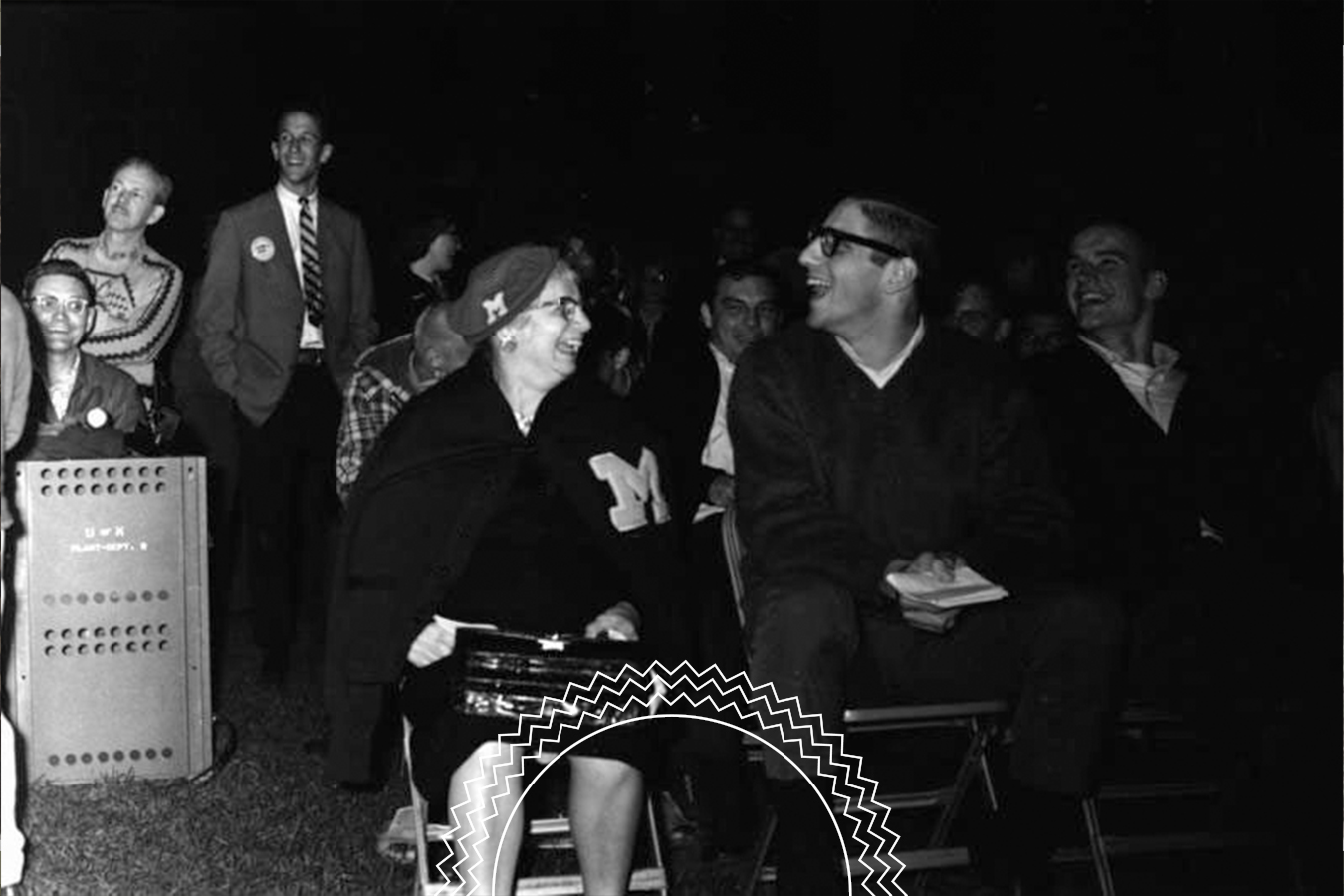 A group of people sitting on chairs outdoors at night, some smiling and laughing, with a machine visible in the background.