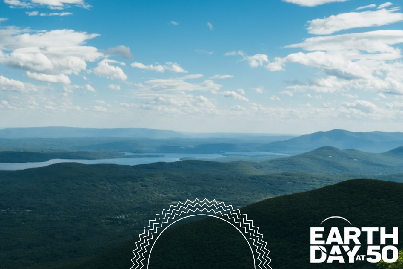 View of rolling green hills and distant lakes under a blue sky with clouds, featuring the “Earth Day at 50” logo in the lower right corner.