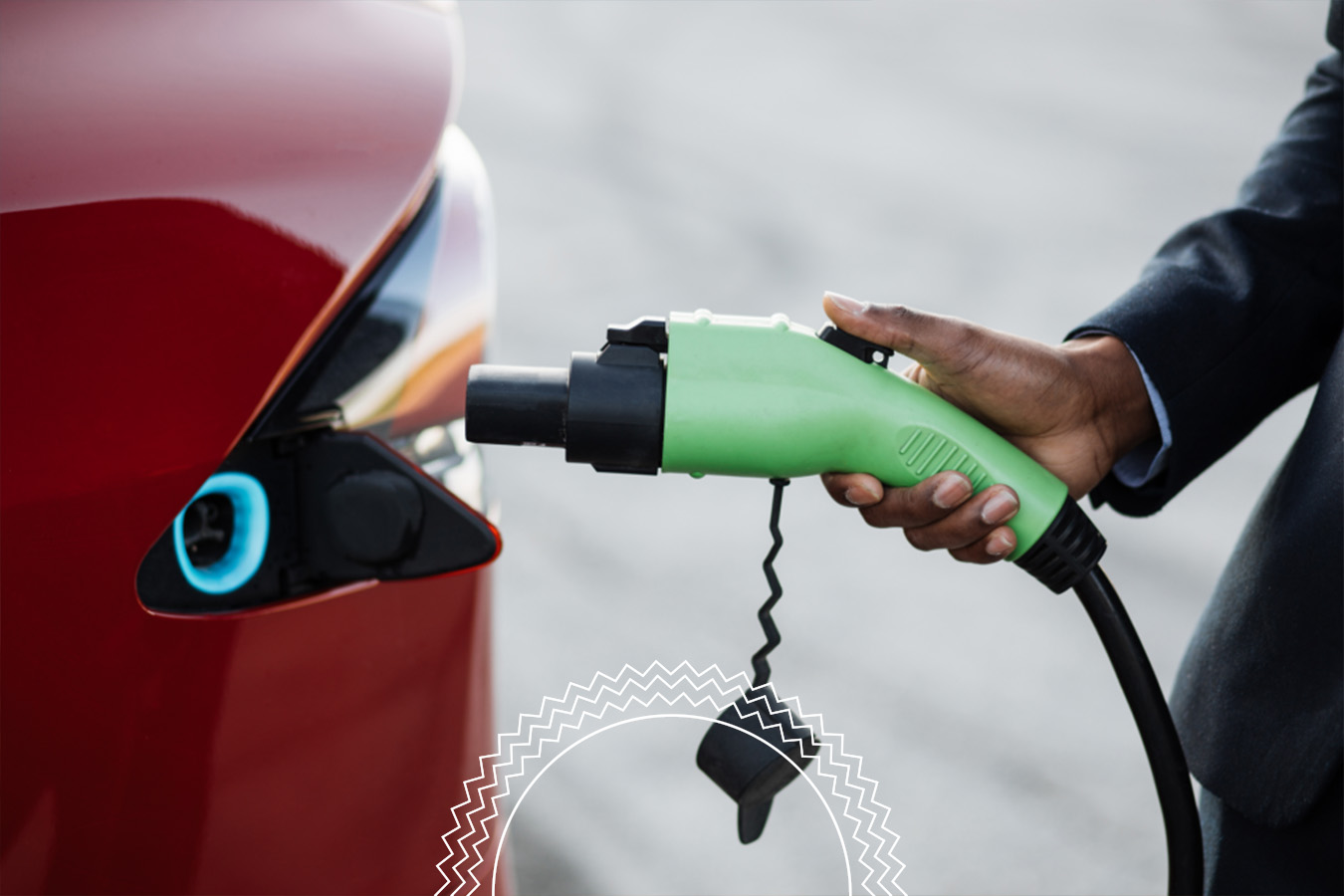A person in a suit plugs a green charging cable into a red electric car at a charging station.