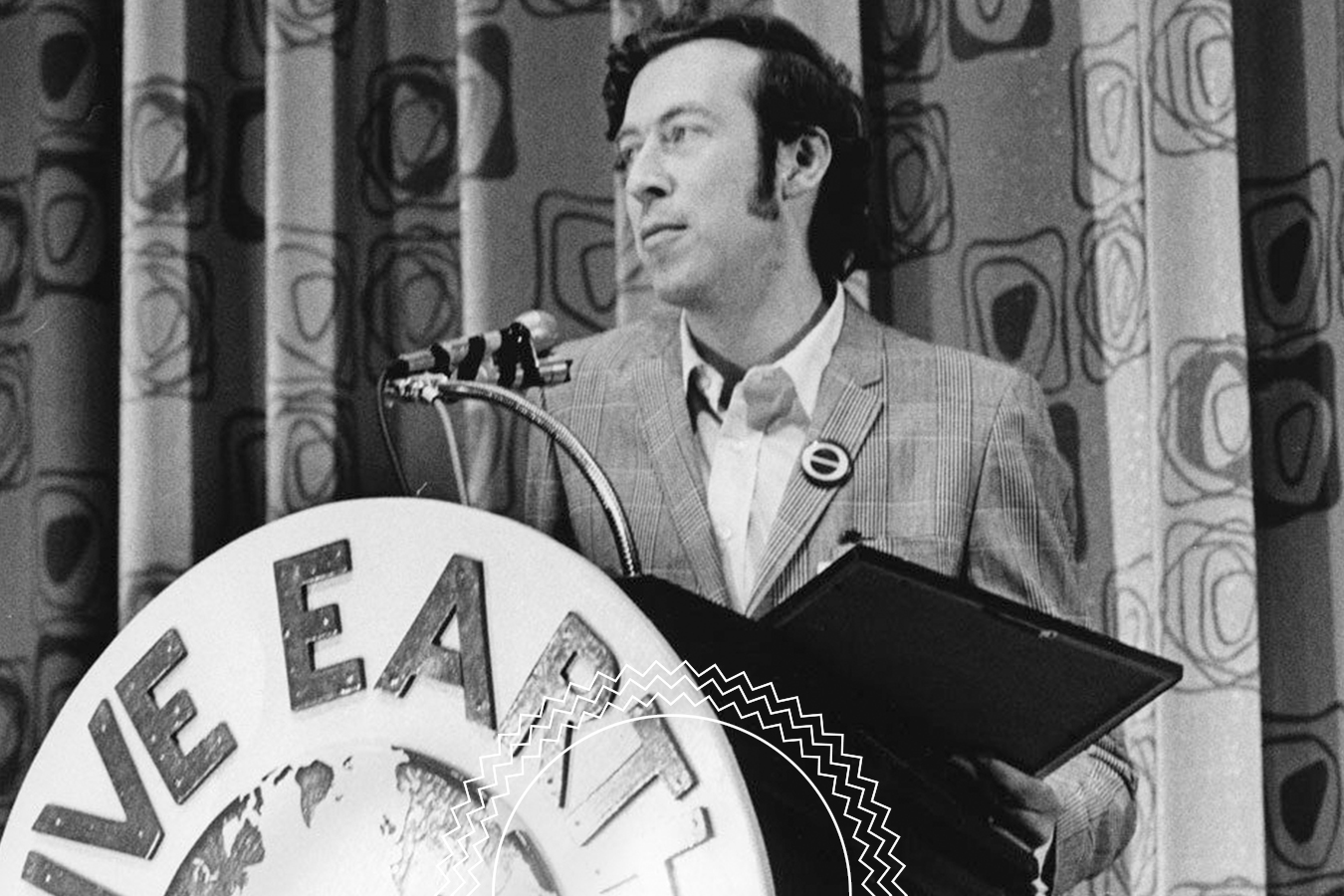 A man in a plaid suit speaks at a podium with a “LOVE EAR” sign, standing in front of a patterned curtain and holding a folder.