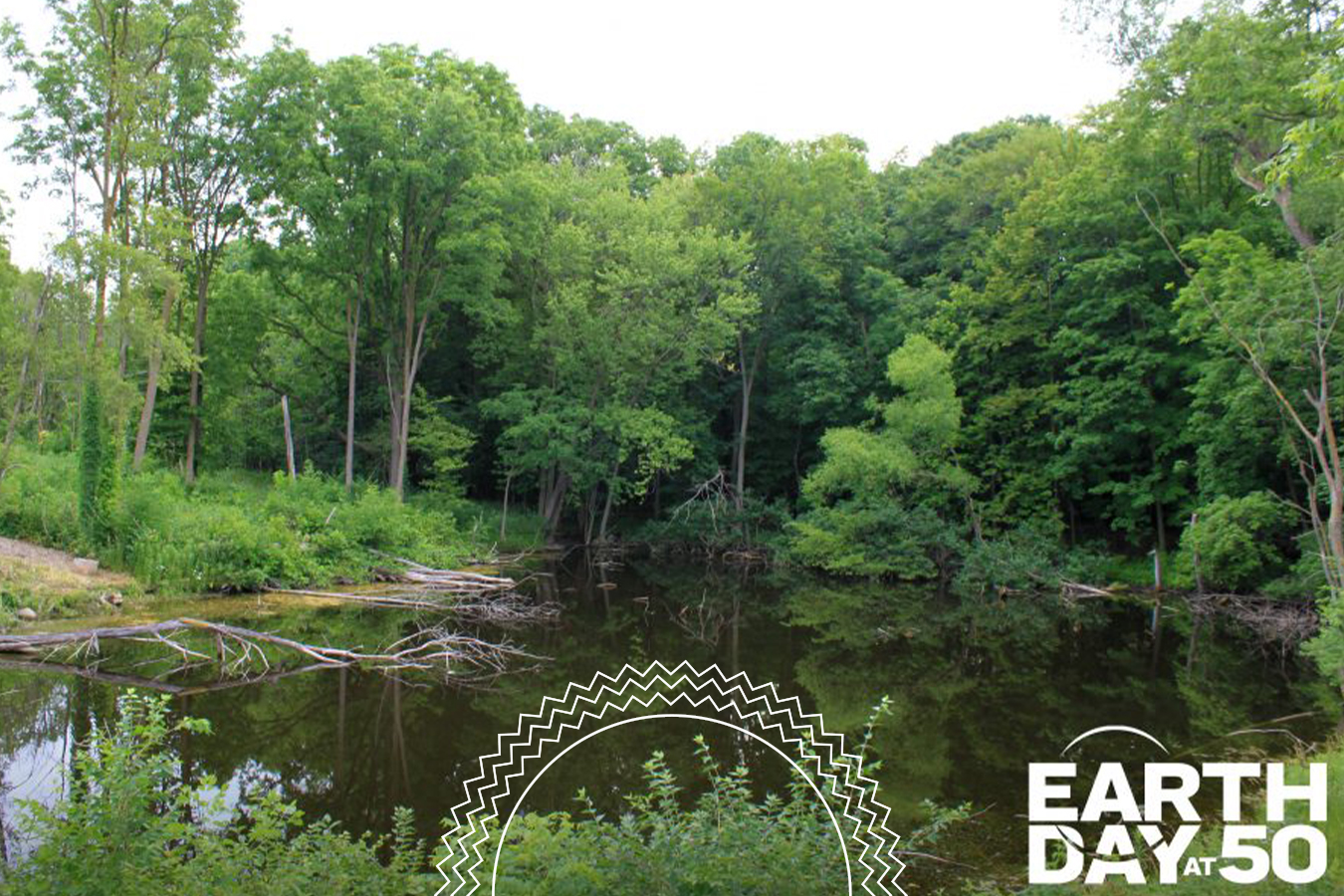 A forested pond with surrounding green trees is shown. The “Earth Day at 50” logo is visible in the lower right corner.