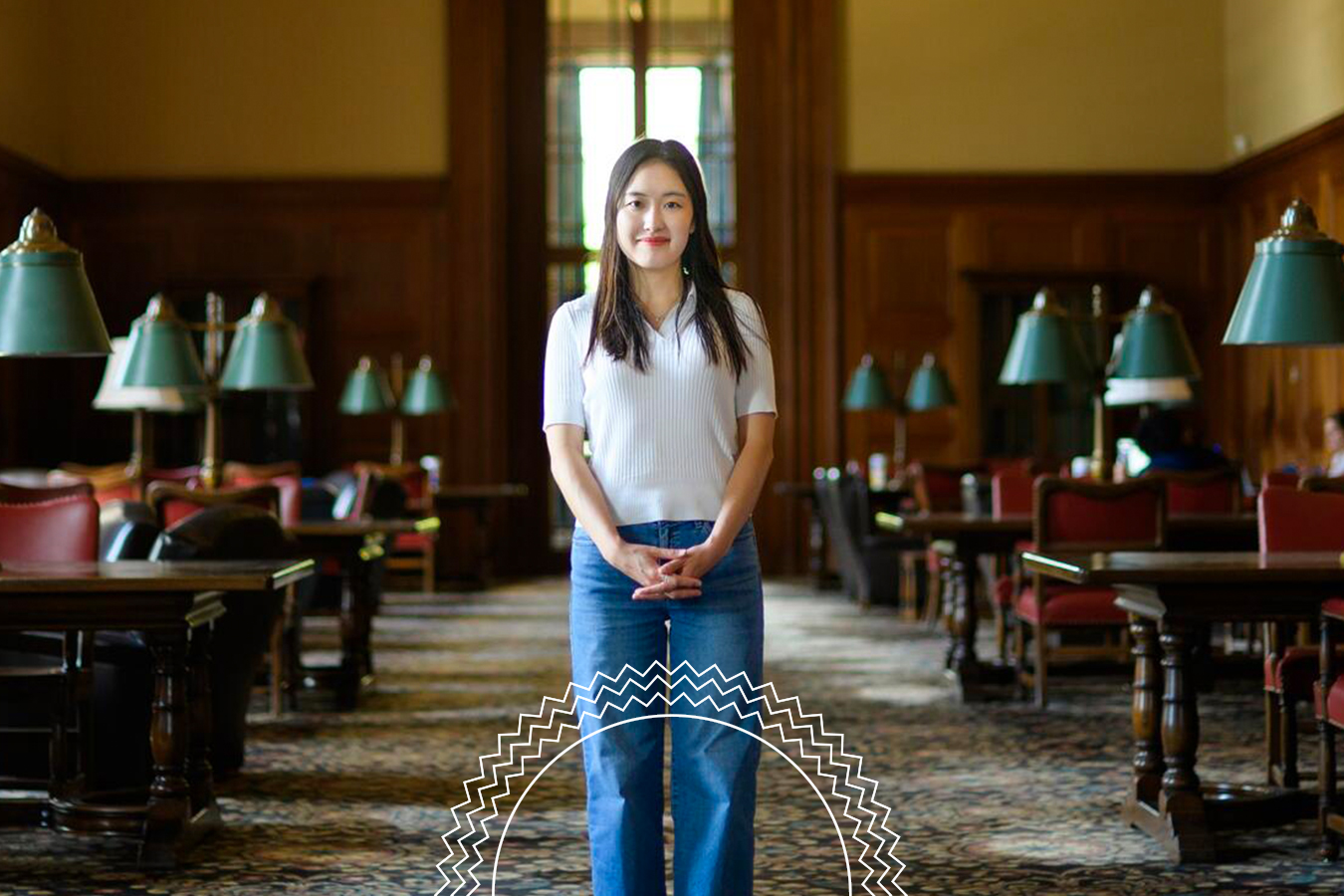 A woman stands in the center of a library reading room with wooden walls, green desk lamps, and large windows in the background.