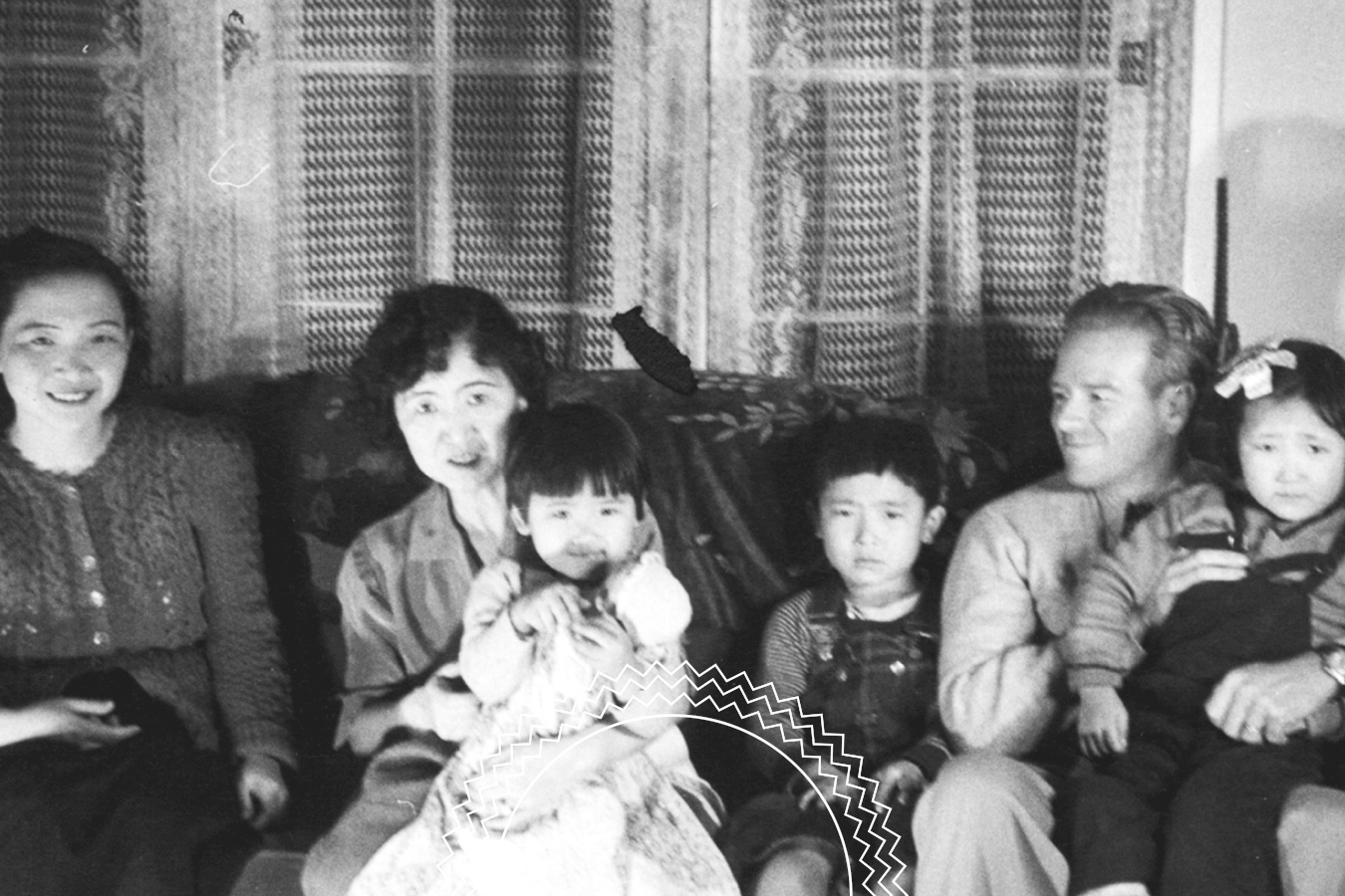 Black and white photo of three adults and three children sitting on a couch in front of a curtained window, all facing the camera.