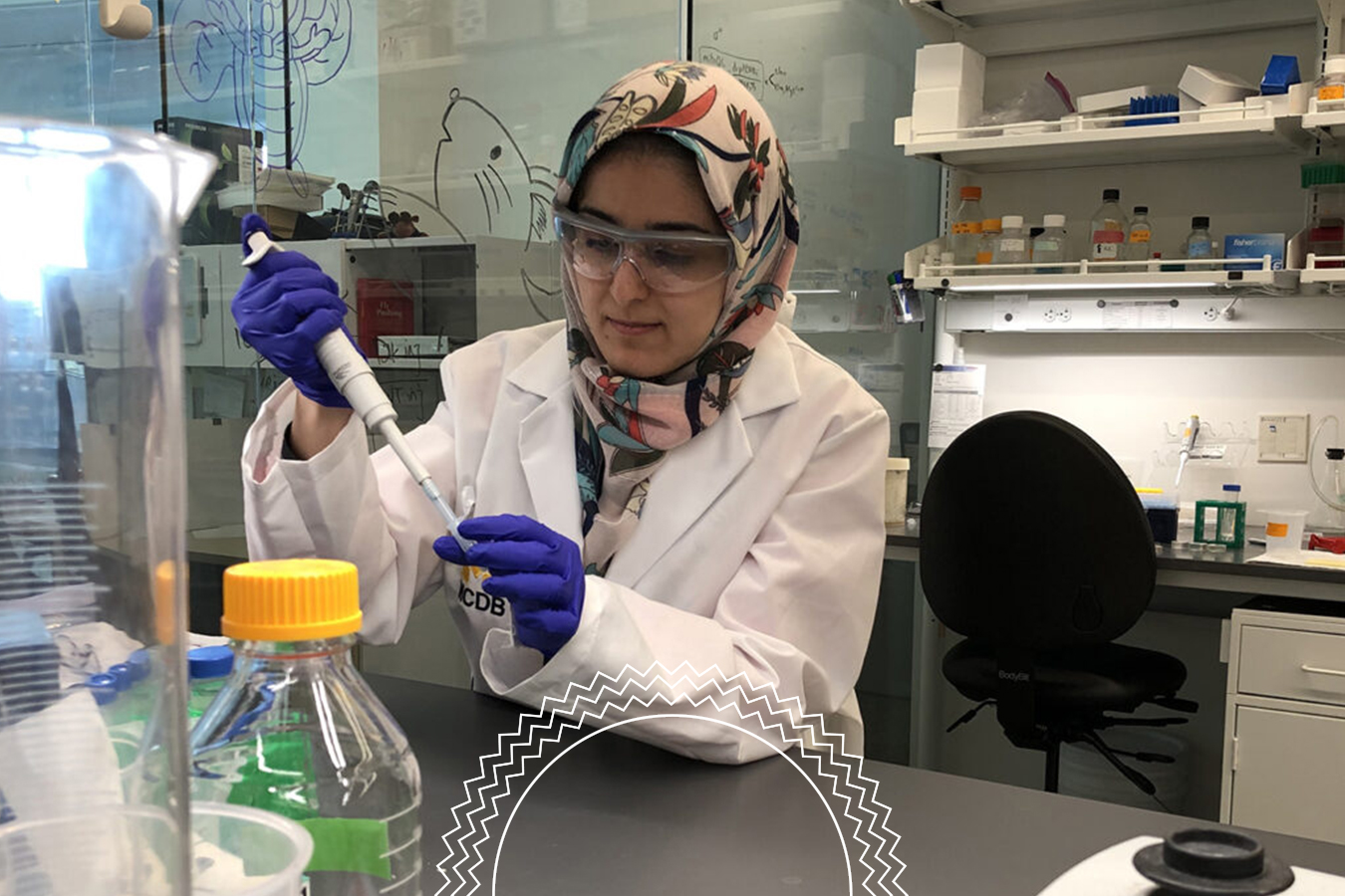 A scientist in a lab coat and gloves uses a pipette at a laboratory bench with bottles, glassware, and scientific equipment nearby.