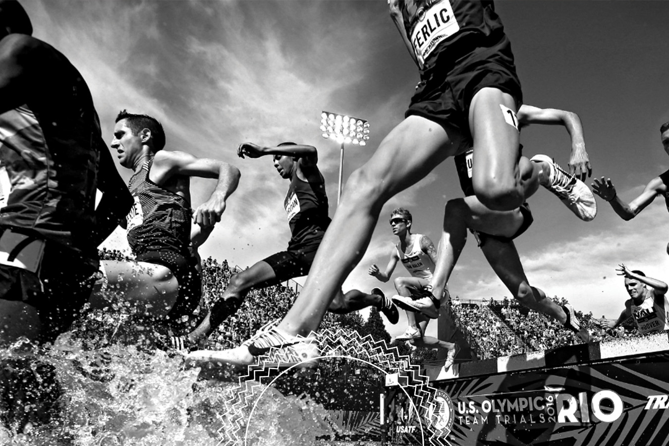 Track athletes leap over a water hurdle during a steeplechase race at the U.S. Olympic Team Trials, with a crowd and stadium lights in the background.