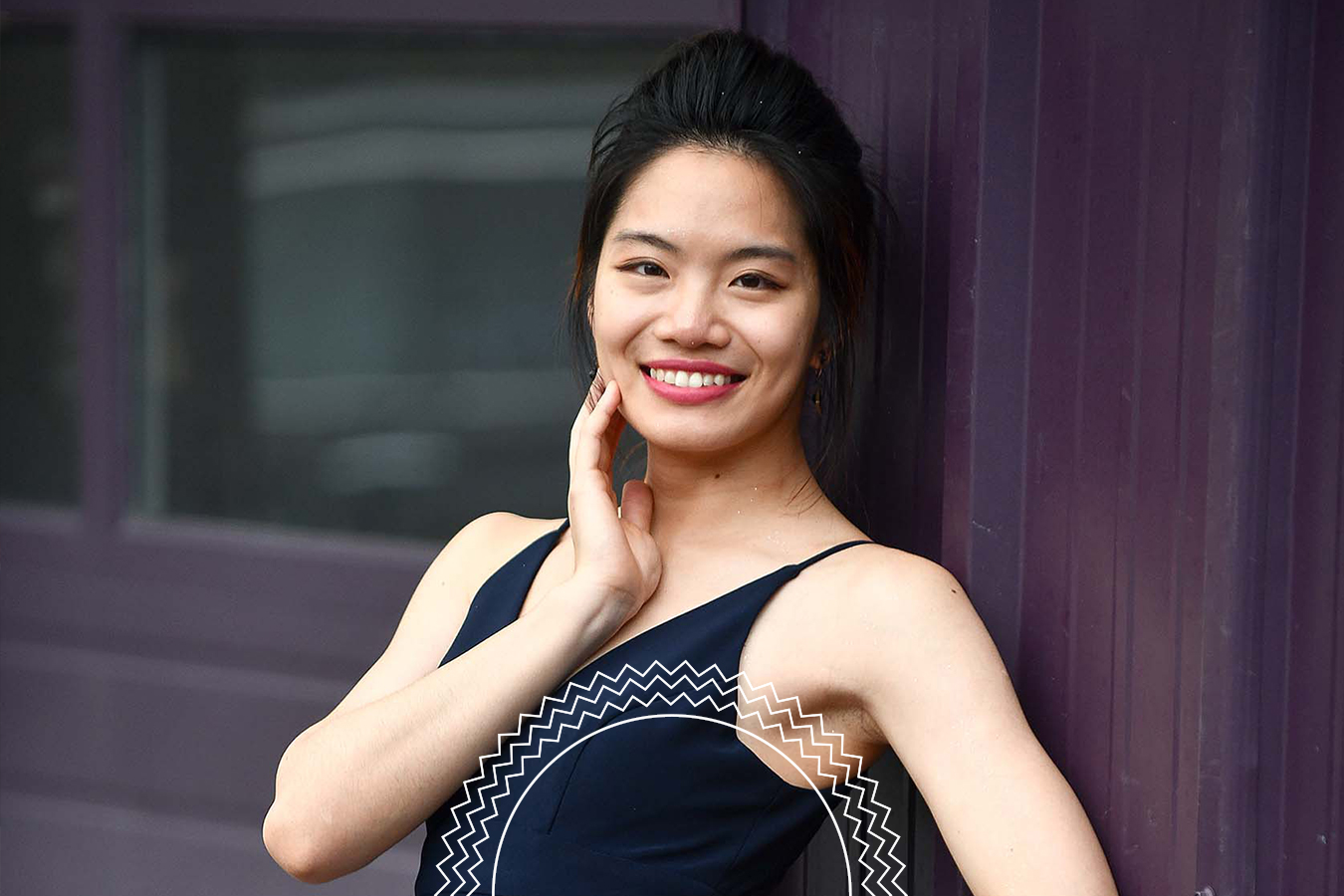 A woman wearing a dark sleeveless top stands against a purple wall, smiling at the camera with one hand touching her face.