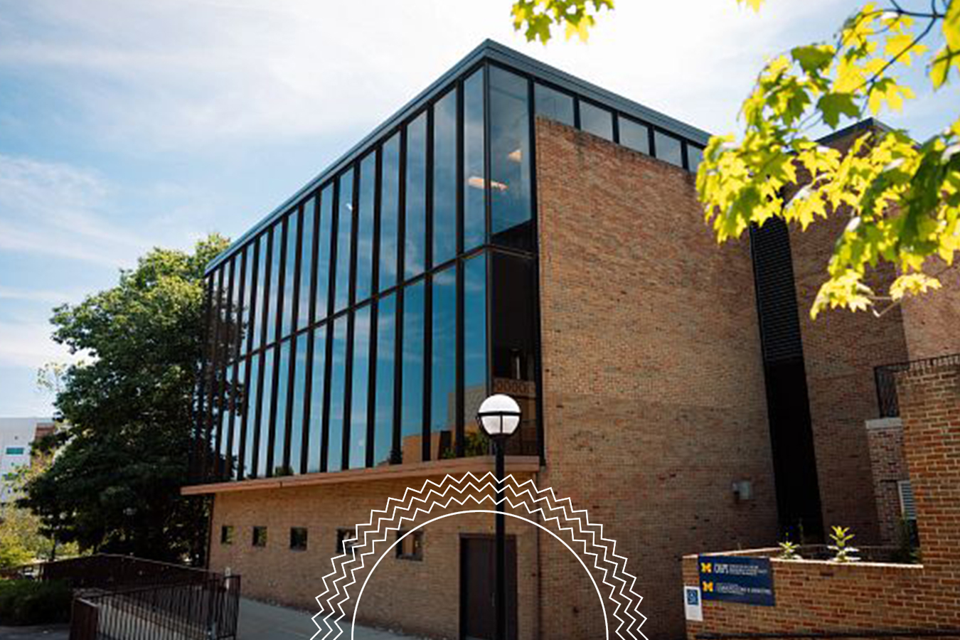 A modern brick building with large glass windows, surrounded by trees, under a clear sky; a University of Michigan sign is visible near the entrance.