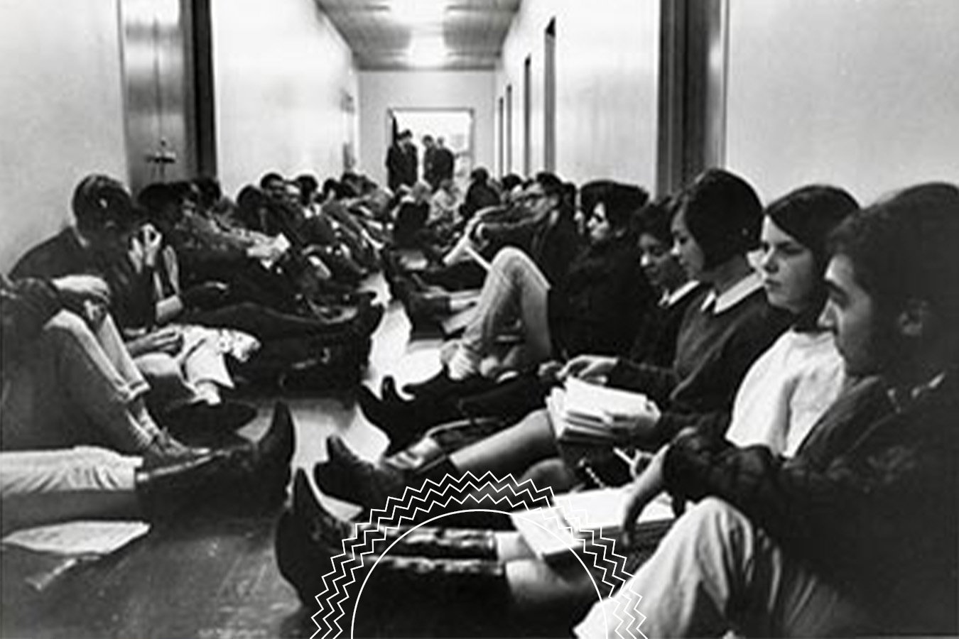 Black and white photo of people sitting on the floor in a hallway, facing each other in two rows, appearing to wait or participate in a group activity.