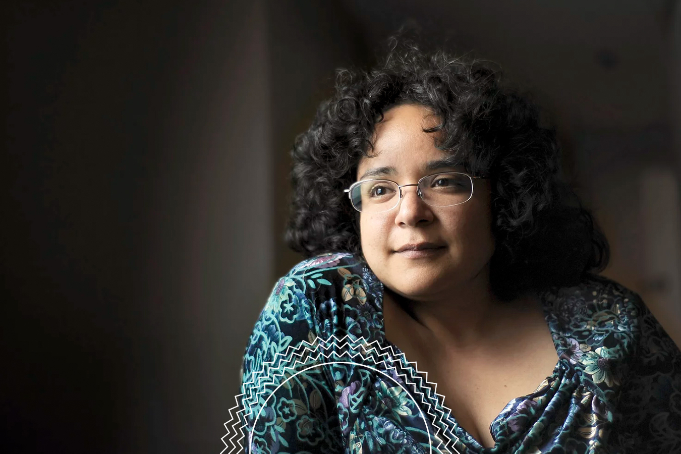 A person with curly dark hair and glasses, wearing a blue patterned top, looks thoughtfully out of frame against a dimly lit background.