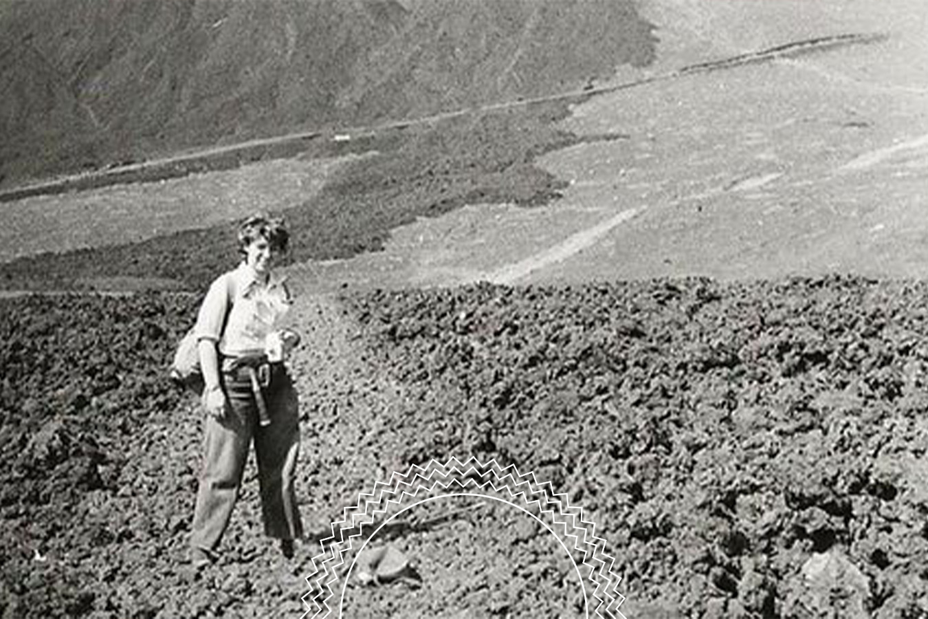Person standing on rough, rocky terrain with mountains in the background, wearing a shirt and pants, holding an object in one hand.