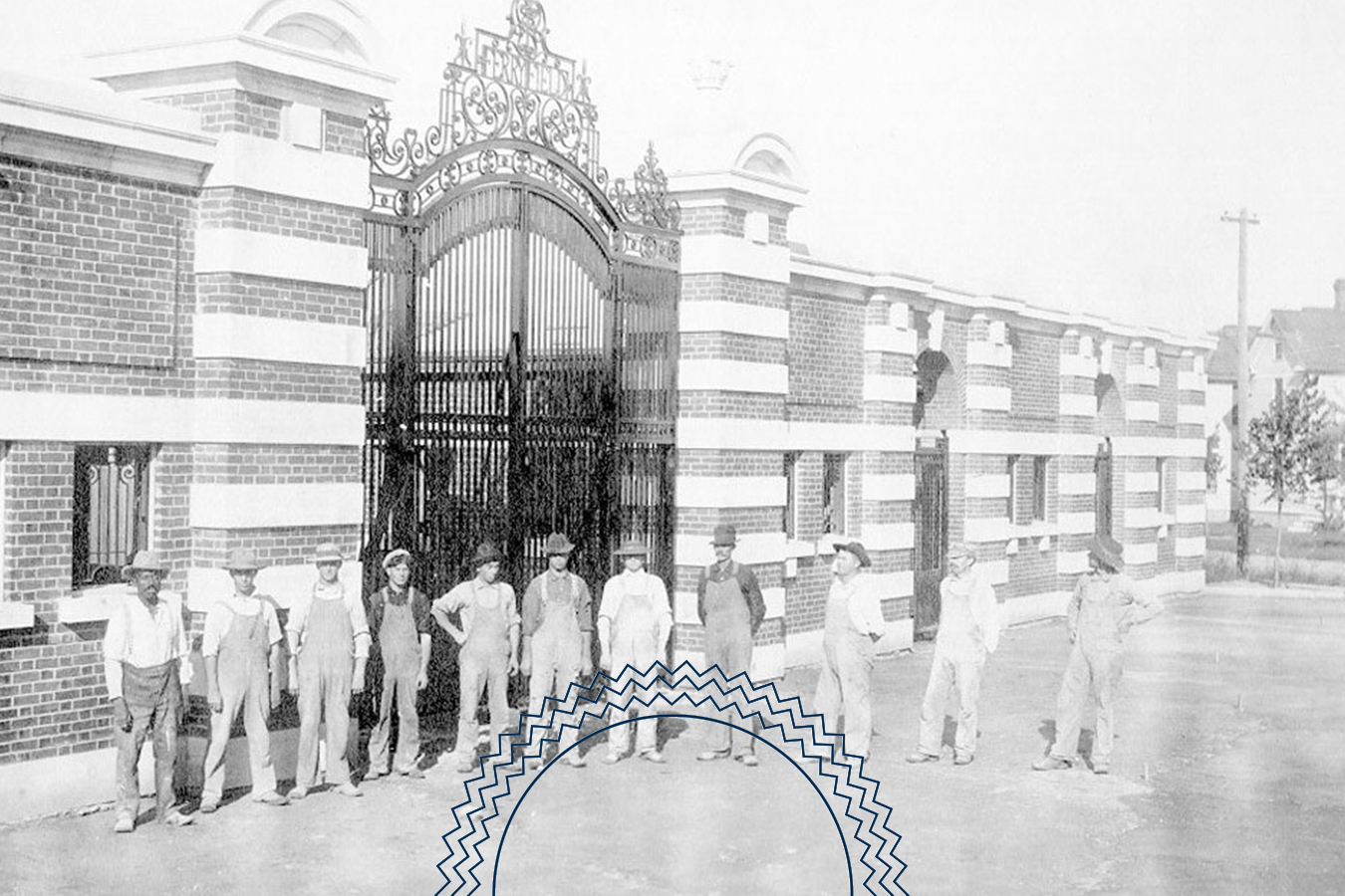 A group of men stand in front of an ornate iron gate and brick building, posing for a photo on a paved surface in daylight.