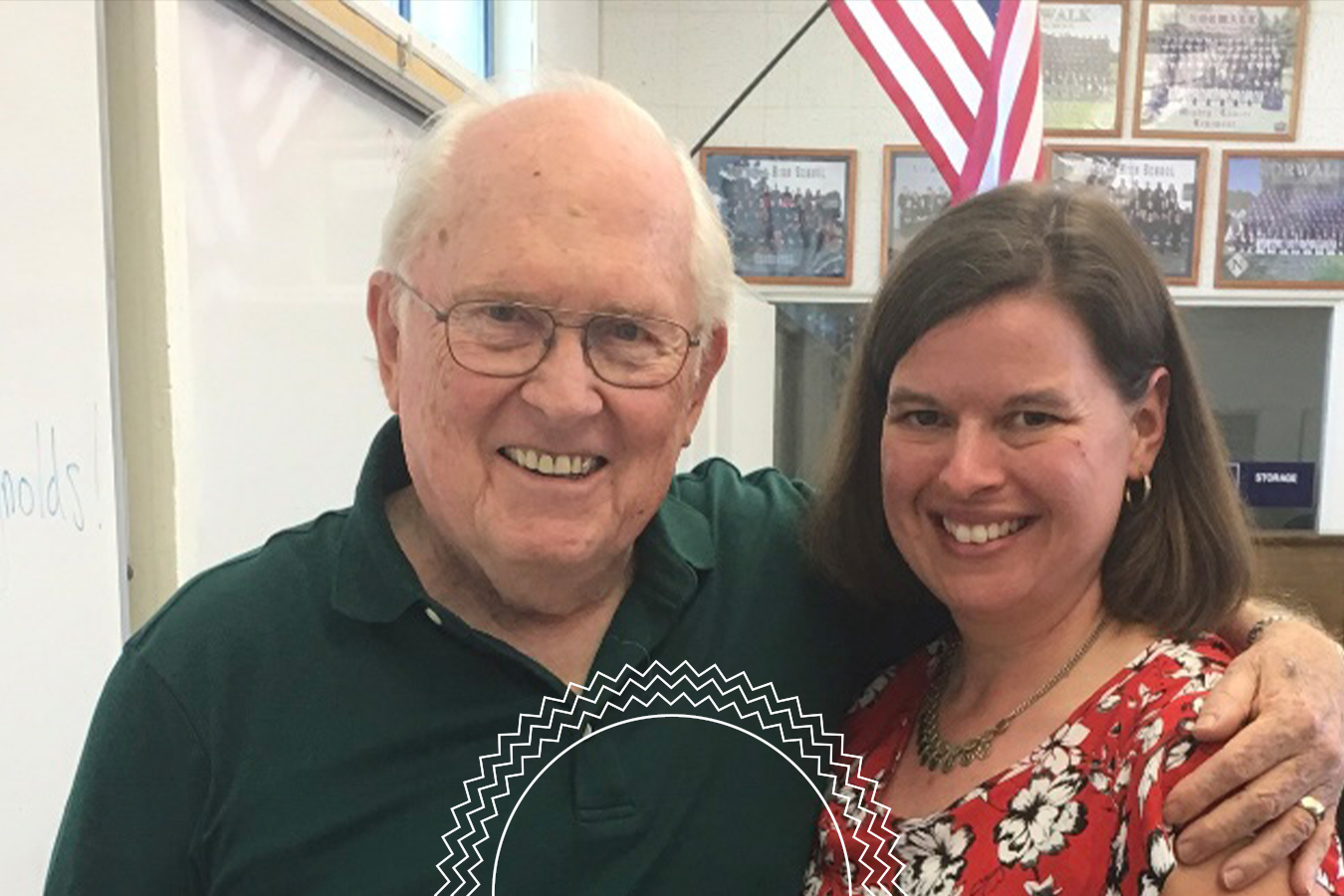 An older man and a woman stand together smiling in a classroom, with an American flag and framed photos on the wall in the background.