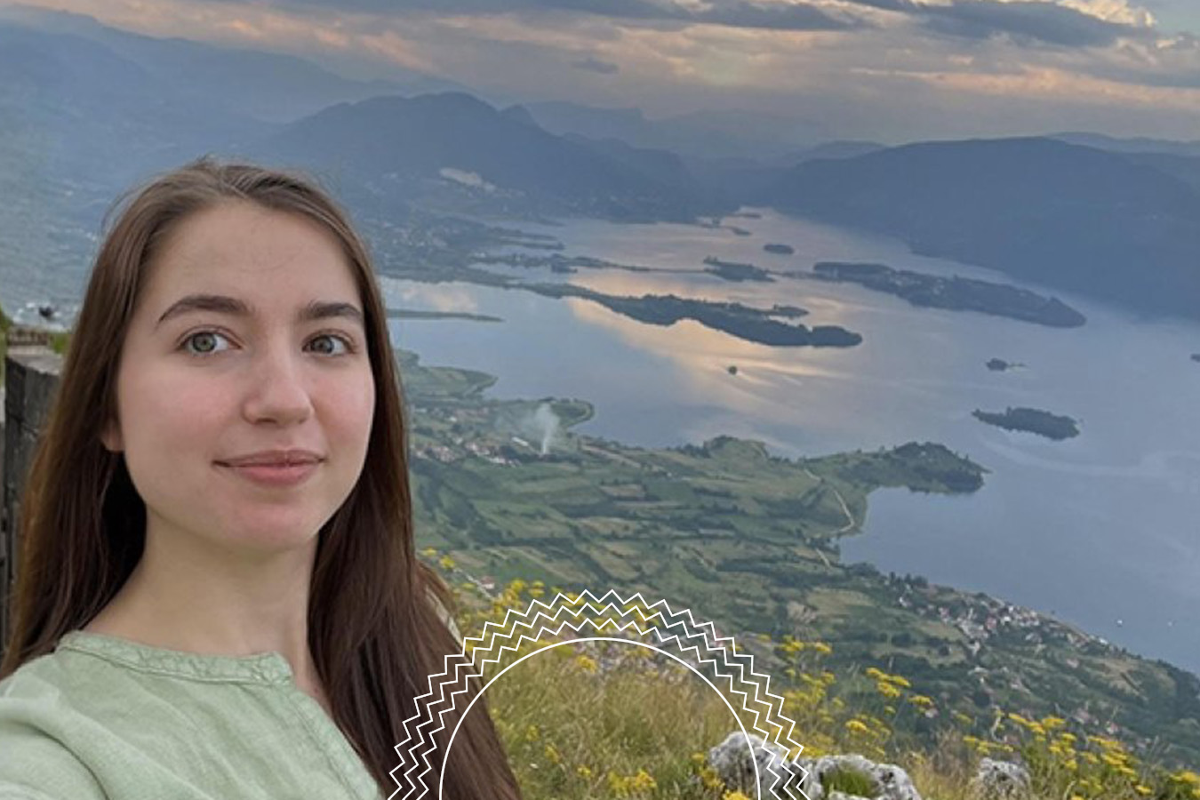 Woman taking a selfie on a hill with a scenic view of a lake, islands, and mountains in the background under a cloudy sky.