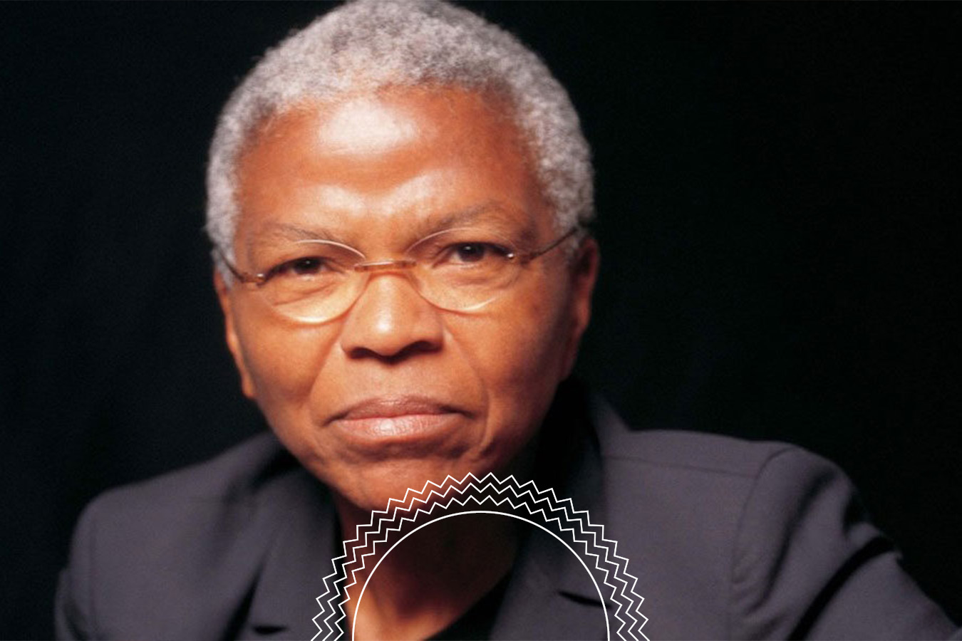 An older person with short gray hair, glasses, and a dark jacket looks directly at the camera against a black background.
