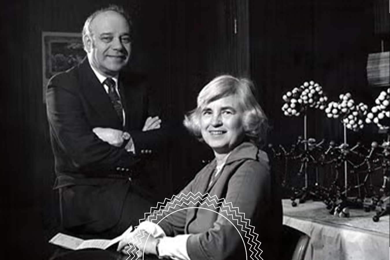 A man stands beside a seated woman in a room with molecular models on a table in the background. Both are dressed in formal attire and facing the camera.