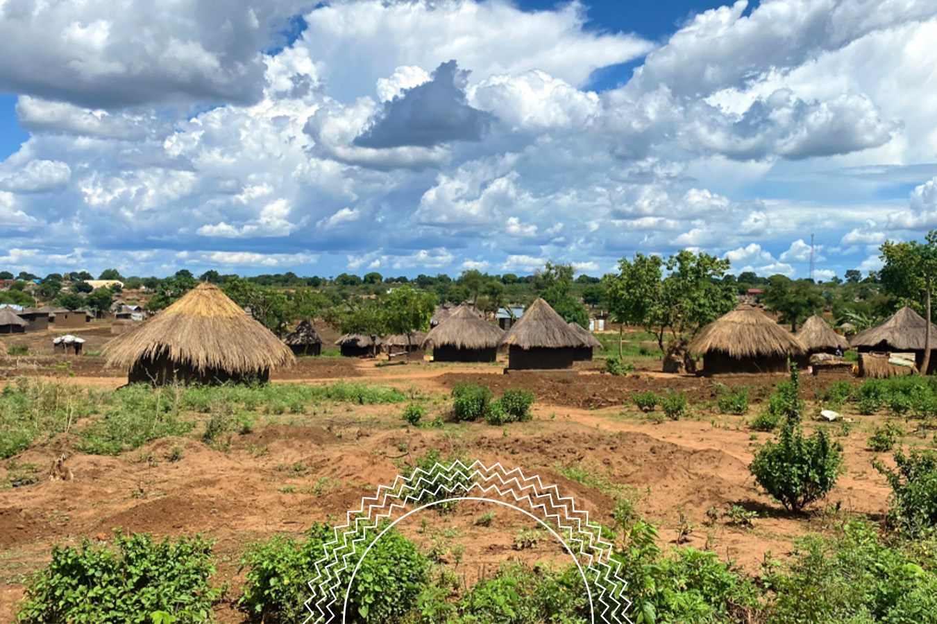 Thatched-roof huts are scattered across a rural landscape with red soil, green bushes, and a cloudy blue sky in the background.