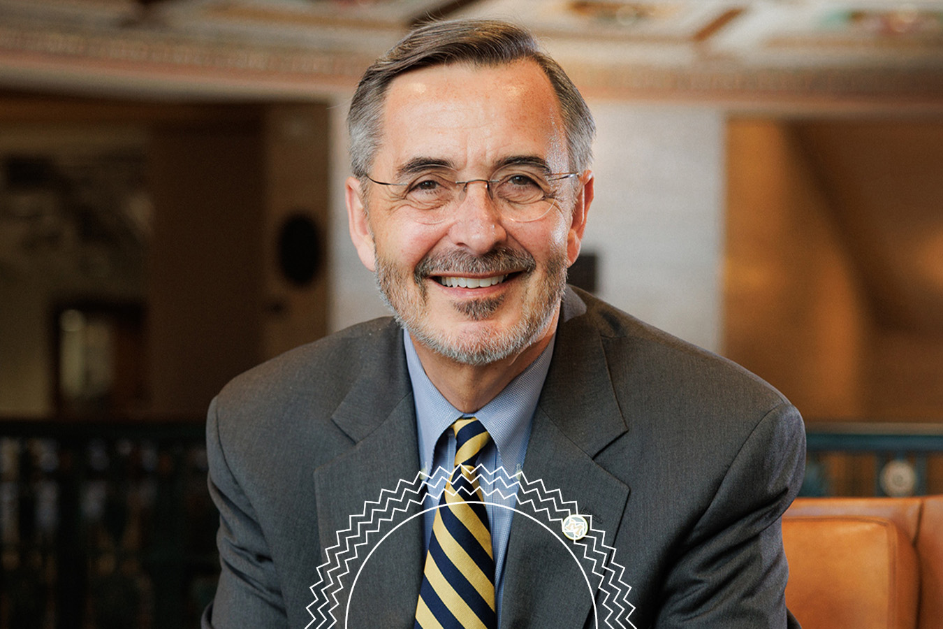A man in a gray suit, striped tie, and glasses sits indoors, smiling at the camera. The background is softly blurred, with architectural details visible.